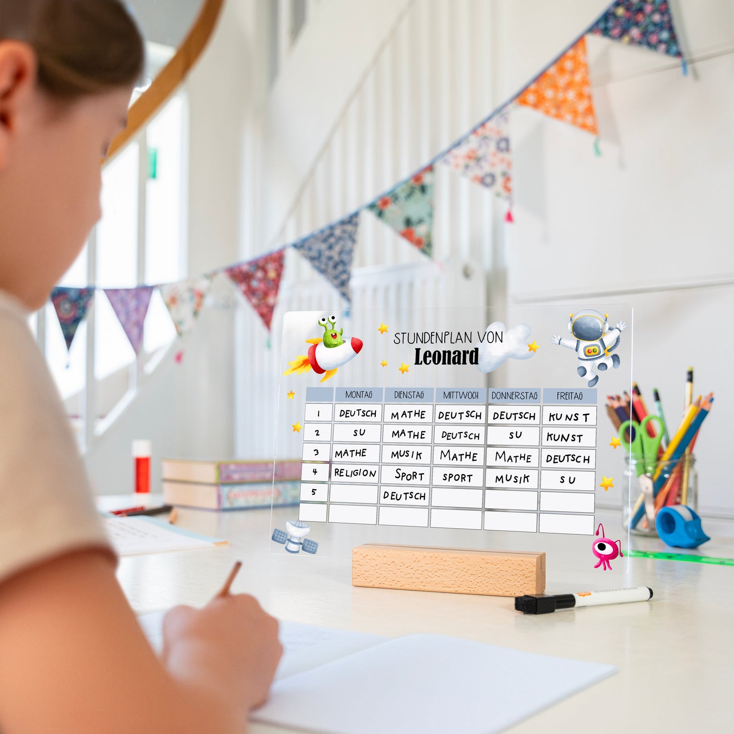 Kind sitzt am Schreibtisch, auf dem Tisch steht der personalisierte Stundenplan Astronaut 1 aus Acryl mit Holzsockel und Stift